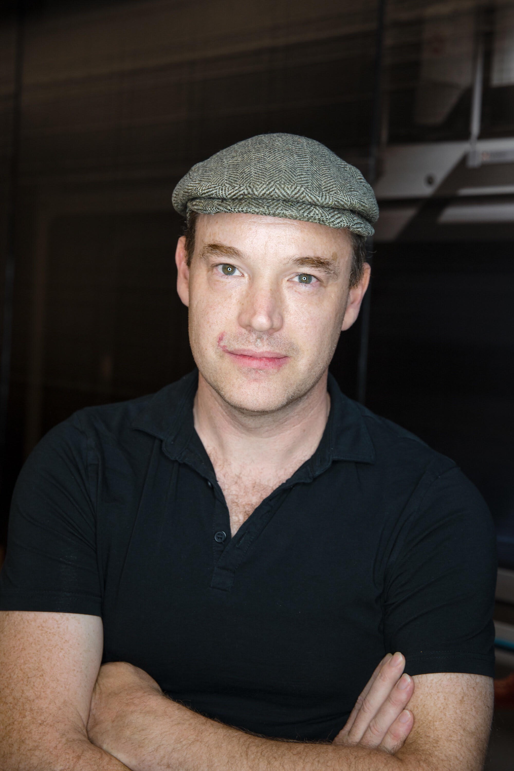An upper-body shot of Owen. He has dark hair and blue eyes and is wearing a grey beret and a dark polo shirt. His arms are crossed.