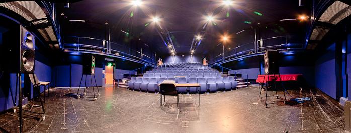 A panoramic view of the Rymer auditorium from the stage. The walls are blue, the floor is dark brown. There are 3 large loudspeakers visible with a table in the centre of the stage. Lines of blue fold-down chairs can be seen in tiers going towards the back of the room with steps up each side.